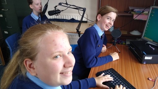 Three students at a desk, one sitting in front of a recording microphone. They are all looking towards the camera smiling.