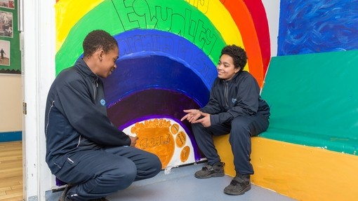 Two students sitting in front of a rainbow painted on a wall that says equality 