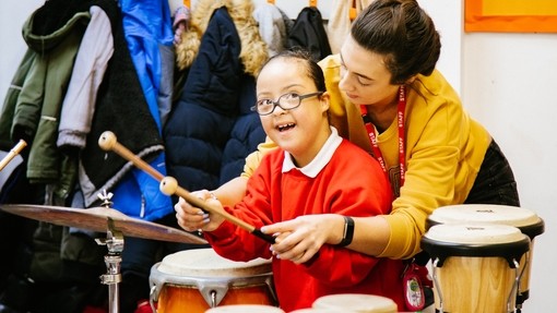 Young primary pupil being taught to play bongos by her teacher who is standing behind her