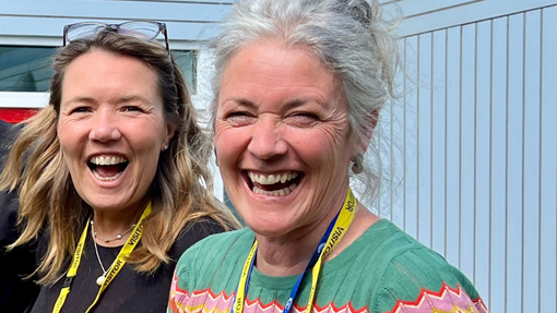 A woman with grey hair and a bright green top is looking to the camera, smiling. Another woman is smiling behind her.