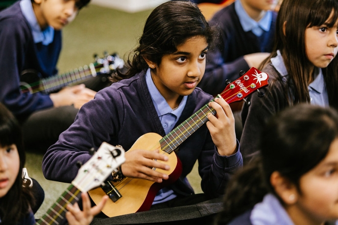 A young Asian girl sits cross legged playing a ukelele, looking up and away from camera