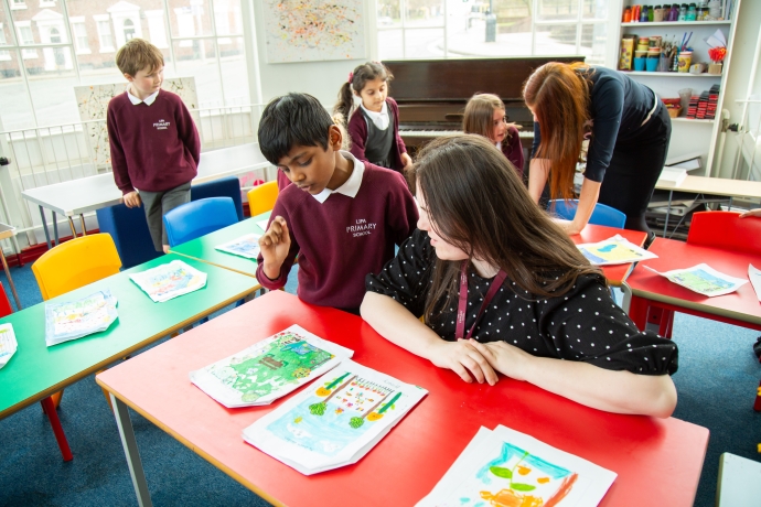 Students looking closely at drawings and designs on the table with a teacher sat on the table at the front and a teacher leaning over a table in the back righthand corner 