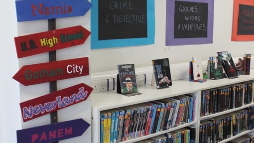 A display of a signpost with different coloured signs pointing in different directions, with books shelves on the right 