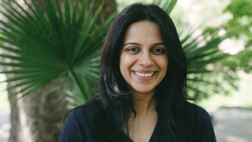 Head shot of a smiling British Asian woman with a black top on and standing in front of a tree 