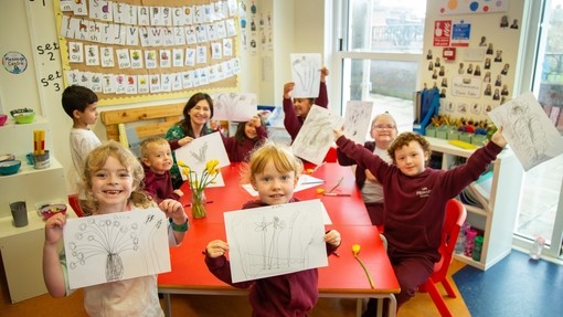 Children in a classroom standing up around the table showing off their drawings