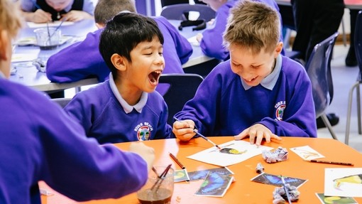 An image of two children sat at a table, one is laughing and the second has his head down whilst concentrating on painting. 
