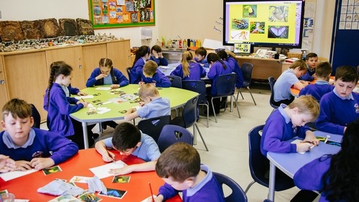An image of a classroom with around 20 students sat on circular desks, concentrating on painting an image of a bird. 