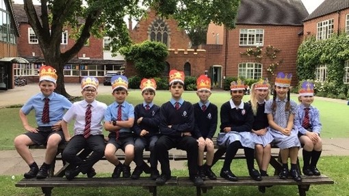 An image of a line of pupils sat on a bench outside, wearing homemade crowns. 