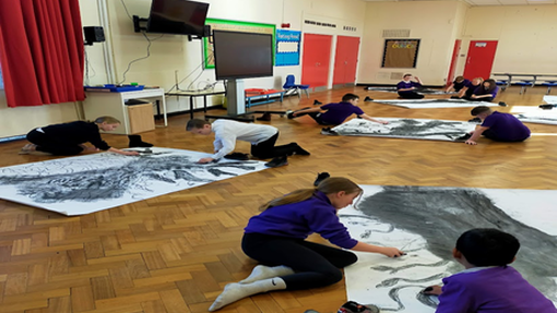 Children wearing purple jumpers sit on the floor of a school hall while drawing on large pieces of paper