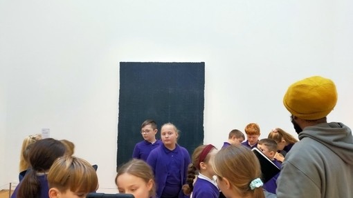 Children wearing purple jumpers and a man wearing a yellow beanie hat and grey hoodie stand in an art gallery with while walls.