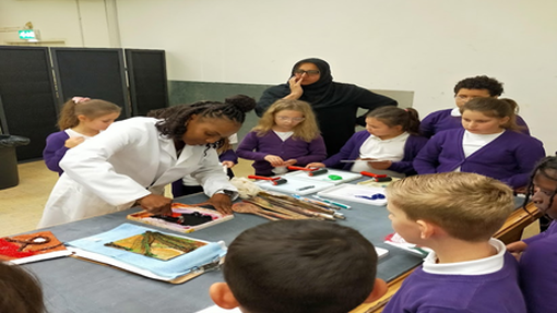 Children wearing purple jumpers and a woman wearing a black headscarf watch a woman wearing a white coat demonstrate how to use an object to do print painting.