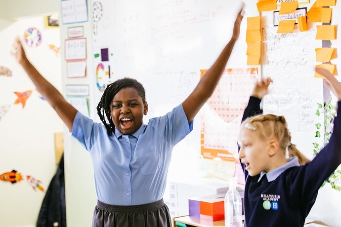 A young black girl dancing with her arms raised in front of maths-related charts and posters on the wall. A Caucasian girl with blond hair is to her right with arms raised 