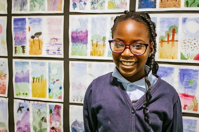 A young black girl with glasses stands in front of her art work on display, smiling proudly