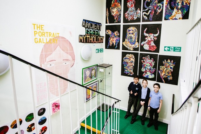 Camera looks down a stairwell that is displaying young people's art work from floor to ceiling with two pupils standing halfway down the stairs