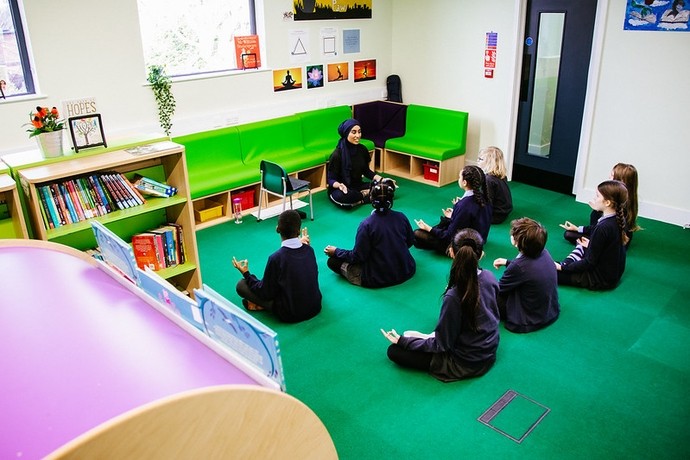 A class of primary pupils with their backs to the camera are seated cross-legged on the floor of a school library in a yoga lesson