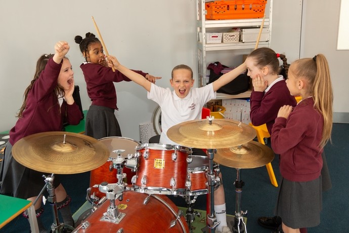 A young female pupil sat at a drum kit with her arms raised and holding a rock star pose, while her classmates make silly faces at her, stood around her in a semi circle 
