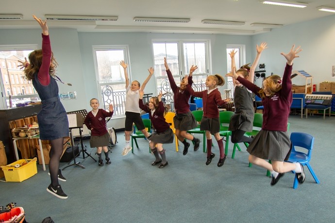 Pupils in a music room with their teacher, all jumping up with arms in the air