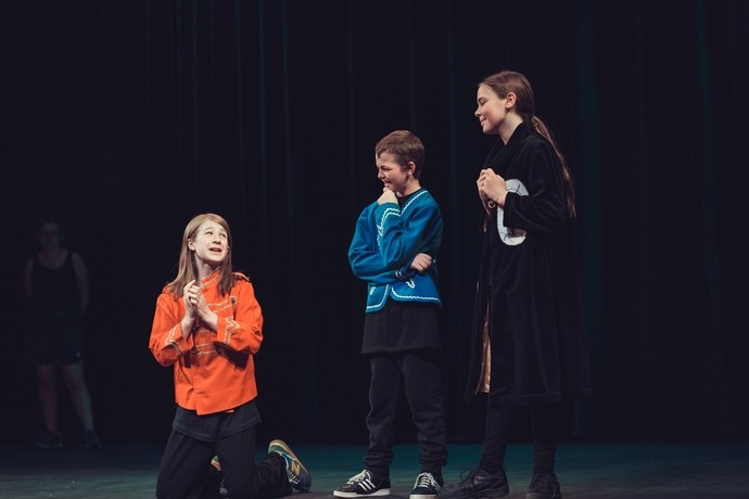 Three young people on stage performing - one kneels, the other two stand next to her