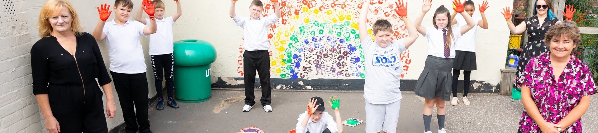 Students and teachers standing against a wall mural of a rainbow they painted with their hands