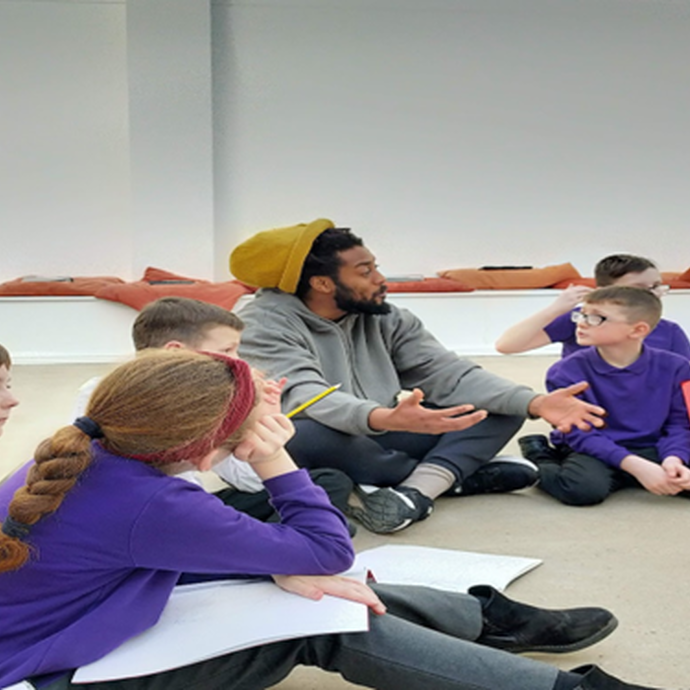 Children in purple jumpers sit on the floor around a man wearing a yellow beanie hat.